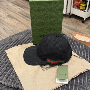 Svart Gucci keps med GG-mönster - Svart keps från Gucci med klassiskt GG-monogram över hela ytan. Kepsen har en böjd skärm och justerbar rem baktill. På sidan finns Guccis ikoniska röd-gröna rand. Materialet är bomull och kepsen har en snygg, mörk look som sticker ut.
