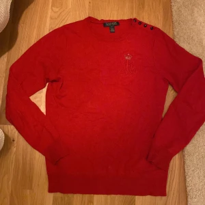 Röd stickad tröja från Ralph Lauren - Töd stickad tröja från Ralph Lauren med broderad RL-logo och krona på bröstet. Tröjan har rund hals, långa ärmar och dekorativa knappar på axeln. Tröjan har lite fläckar framtill men som säkert går bort i tvätten, inget som man lägger märke till. Storleken är large men jag uppfattar den snarare som en Medium. Materialet: 85% silk & 15% cashmere