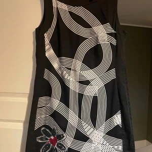 Svartvit kort Desigual klänning med grafiskt mönster - Cool svart ärmlös klänning från Desigual med grafiskt vitt mönster och en broderad blomma med rött hjärta nertill. Klänningen är helfodrad o har dragkedja bak och är i en rak, kort modell som känns både modern och edgy.