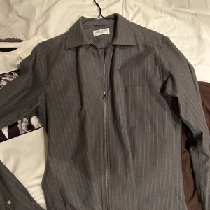 Grå randig overshirt från John Herrick - Snygg grå overshirt från John Herrick med tunna vita ränder och dragkedja framtill. Klassisk krage och en bröstficka ger en clean look. Perfekt att slänga över en t-shirt för en chill och stilren vibe. Har aldrig använts 