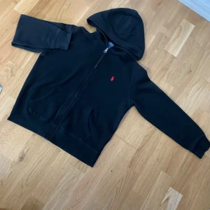 Svart hoodie från Polo Ralph Lauren - Svart hoodie från Polo Ralph Lauren med röd broderad logga på bröstet. Tröjan har huva, dragkedja framtill och två fickor. Tillverkad i mjuk bomull och polyesterblandning, perfekt för chill dagar. Klassisk och stilren design som alltid funkar.