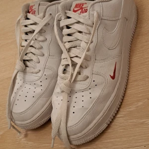 Nike Air Force 1 vit med röd swoosh - Nike Air Force 1 sneakers i vitt skinn med röda detaljer på swoosh och logga. Klassisk låg modell med rund tå, perforerad tåbox och platt sula. Skorna har snörning och subtila paneler i olika texturer. Perfekt för dig som gillar clean och stilren streetwear.