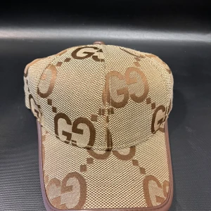 Mönstrig beige keps - Snygg beige keps med det klassiska mönstret i brunt över hela kepsen. Tillverkad i bomull med detaljer i brunt skinn baktill. Perfekt för dig som vill ha en lyxig och trendig accessoar med tydlig designerstil.