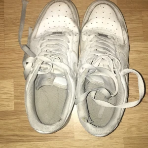 Nike Air Force 1 vita sneakers - Klassiska Nike Air Force 1 sneakers i helvitt skinn med rund tå och platt sula. Skorna har snörning och den ikoniska swoosh-loggan på sidan. Perfekt för dig som gillar streetwear och vill ha en tidlös sneaker i garderoben.