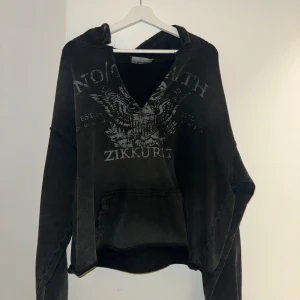 Svart hoodie från No Faith Studios - Svart hoodie från No Faith Studios med coolt örnmotiv och text på bröstet. Tröjan har huva, känguruficka och en rå, lite sliten v-ringad hals. Oversized passform och långa ärmar, perfekt för dig som gillar streetwear.