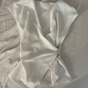 Vit omlottopp med gulddetalj - Snygg vit omlottopp med ärmlös design och v-ringning. Toppen har en cool guldfärgad metallpinne som håller ihop omlotten framtill. Perfekt för dig som gillar stilrena och trendiga plagg med en twist.