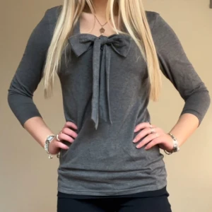 Grå topp med rosett och trekvartsärm - Snygg grå topp med stor rosett framtill och trekvartsärmar. Tröjan har även krage