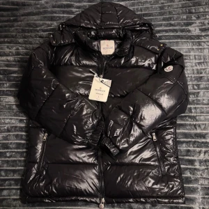 Moncler jacka - Säljer en svart, glansig pufferjacka från Moncler med huva och dragkedja framtill. Jackan har två sidofickor med dragkedja och ikonisk Moncler-logga på ärmen. Insidan har ett coolt serietidningsmärke. Perfekt för kalla dagar och riktigt snygg streetstil.