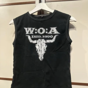 Svart W:O:A linnetopp med tryck - Cool svart ärmlös linnetopp med vitt W:O:A-tryck och en stor tjurskalle på bröstet. Rund halsringning och kontrastsömmar längs kanterna. Perfekt för dig som gillar festivalstil och rockvibe.