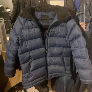 Blå Peak Performance dunjacka M - Snygg blå pufferjacka från Peak Performance i storlek M. Jackan har huva, två sidofickor med dragkedja och ett stilrent broderat logotyp på bröstet. Perfekt för kalla dagar med sin fluffiga och quiltade design. Materialet är syntet och dun.