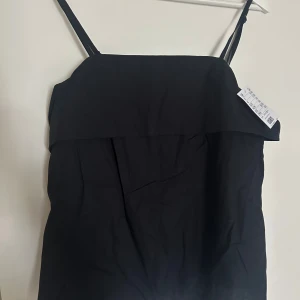 Svart linne med smala axelband - Fint svart linne med vikt kant upptill och smala justerbara axelband. Dragkedja i ryggen, helt ny med prislapp kvar! 