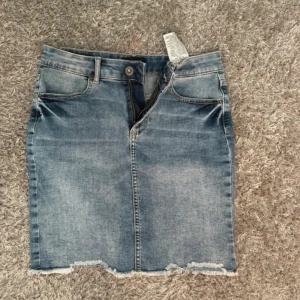 Blå jeanskjol med fransad kant - Säljer en blå jeanskjol med fransad kant och klassiska fickor både fram och bak. Kjolen har bälteshällor, knapp och dragkedja framtill. Den är i en kort modell och har en snygg tvättad look som ger en chill vibe.