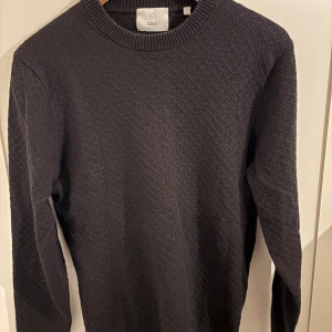 Svart stickad tröja från Bläck - Säljer en svart stickad tröja från Bläck i storlek M. Tröjan har ett diskret mönster i tyget och rund halsringning med ribbade muddar. Perfekt för lager-på-lager.