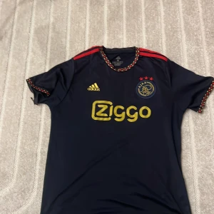 Ajax x Adidas mörkblå fotbollströja M - Ajax fotbollströja från Adidas i mörkblått med röda och gula detaljer. Tröjan har korta ärmar, Ajax klubbmärke på bröstet, tre röda stjärnor och Ziggo-logga i gult. Tillverkad i lätt och ventilerande Aeroready-material.