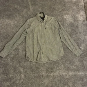 Rutig skjorta från Chevalier M - Snygg rutig skjorta från Chevalier i klassisk passform. Skjortan har grönt och beige rutmönster, knappar med Chevalier-logga och en bröstficka. Tillverkad i mjuk bomull, perfekt för dig som gillar stilren och tidlös design.