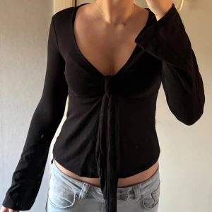 Svart topp med knyt - Säljer en svart topp med lång ärm och djup v-ringning. Toppen har knyt framtill som ger en snygg och trendig look. Croppad passform som visar midjan och mjukt material som sitter skönt mot kroppen. Perfekt för dig som gillar stilrena och coola plagg.