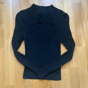 Svart ribbad polotröja med cut out - Snygg svart ribbad polotröja med lång ärm och en cool cut out-detalj framtill. Tröjan har en tight passform och är perfekt för dig som gillar stilrena men ändå unika plagg. Passar lika bra till jeans som till kjol.