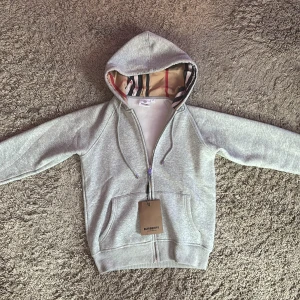 Grå hoodie från Burberry med dragkedja - Snygg grå hoodie från Burberry med klassiskt rutigt foder i huvan. Tröjan har dragkedja framtill, snörning i huvan och två stora fickor. Perfekt för dig som vill ha en stilren och lyxig vardagslook.