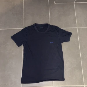 Svart BOSS t-shirt i bomull - Snygg svart t-shirt från BOSS med diskret blå logga på bröstet. Klassisk rund halsringning och korta ärmar. Tillverkad i mjuk bomull som känns skön mot huden. Perfekt för en clean och stilren look.