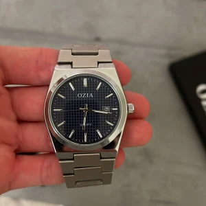 Silverfärgad klocka från OZIA - Snygg klocka från OZIA med silverfärgat armband i metall och mörkblå urtavla med diskret rutmönster. Klockan har datumvisning och stilrena indexmarkeringar. Låset är graverat med OZIA-loggan. Perfekt accessoar för dig som gillar klassisk design. Klockan är vattentät upp till 100m. 