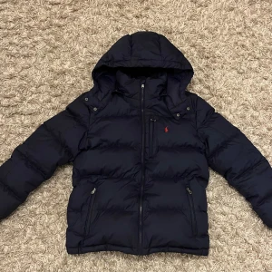 Mörkblå pufferjacka Polo Ralph Lauren XL - Snygg mörkblå pufferjacka från Polo Ralph Lauren med huva som går att ta av. Perfekt för kalla dagar och som är riktigt skön att bära. Jackan är i väldigt fint skick! Junior XL men kan bäras av de som har XS (extra small)