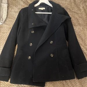 Svart skepparkavaj i ull, strl 36 - Snygg svart skepparkavaj med wdubbelknäppning och bred krage. Jackan är tillverkad i ull och har två fickor framtill samt långa ärmar. Perfekt för dig som gillar klassisk stil med en modern twist.