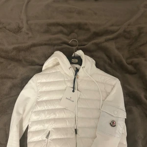 Vit cardigan med huva från Moncler - Säljer en snygg vit pufferjacka från Moncler med huva och snörning. Jackan har quiltad framsida, dragkedja och en ikonisk Moncler-logga på ärmen med ficka. Materialmix av polyester och bomull ger en skön känsla. Perfekt för dig som vill ha en clean och stilren look. Har en pytte liten svart fläck på ryggen men syns knappt och loggan kan skannas med mobil