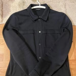 Svart overshirt från Selected Homme - Perfekt svart overshirt från Selected Homme med klassisk krage och tryckknappar framtill. Jackan har  bröstficka med lock och raka ärmar. Perfekt för svalare tider och enkel att matcha med olika outfits.