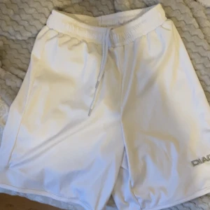 Vita träningsshorts från Diadora - Snygga vita shorts från Diadora med broderad logga i silver på benet. De har elastisk midja med snörning och är tillverkade i ett lätt syntetmaterial som passar perfekt för sport och träning. Modellen är loose fit och har en enkel, stilren design.