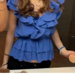 Blå volangblus - Säljer en blå blus med flera lager volanger och omlottdetalj vid halsen. Blusen är ärmlös och har en elastisk midja som ger en snygg siluett. Perfekt för dig som gillar romantiska och trendiga toppar med extra volym. Osäker på storlek eftersom finns ingen lapp