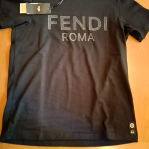 Svart Fendi t-shirt med logga - Svart t-shirt från Fendi med stor FENDI ROMA-logga framtill och en cool detalj med ögonmärke nertill. Klassisk passform och korta ärmar. Tillverkad i mjuk bomull för en skön känsla. Perfekt för dig som vill ha en exklusiv och stilren look. Köptes för typ 1500. Pris kan diskuteras.
