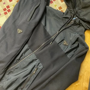 Svart vindjacka från Prada, storlek M - Svart vindjacka från Prada med huva och dragkedja. Jackan har snygga detaljer i form av Prada-logga på bröstet och ärmen, samt flera praktiska fickor med dragkedja. Materialmix av bomull och polyester ger en modern look.