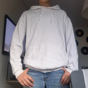 Vit hoodie med huva och ficka - Säljer en stilren vit hoodie med huva och klassisk känguruficka framtill. Tröjan har långa ärmar och ribbade muddar vid ärmslut och nederkant. 