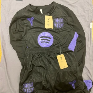 FC Barcelona x Nike x Kobe Bryant player editon - Säljer en svart långärmad FC Barcelona fotbollströja från Nike med lila detaljer och loggor. Tröjan har klubbmärke, Kobe Bryant-logga och Spotify-tryck på bröstet samt lila paneler på sidorna. Materialet är lätt och andas, perfekt för träning eller match.