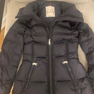 Mörkblå  dunjacka från Moncler - Säljer en svart dunjacka från Moncler med figurnära passform. Jackan har hög krage, tvåvägsdragkedja och två snedställda fickor med dragkedja. Klassisk Moncler-logga på ärmen och quiltad design. Perfekt för kalla dagar. QR koden finns