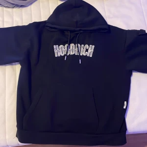 Svart hoodie från Hoodrich med tryck - Svart hoodie från Hoodrich med grön/vit kamouflage broderad logga på bröstet och tryck på ena ärmen. Klassisk känguruficka framtill, snörning i huvan och mjukt material. Perfekt för dig som gillar streetwear och vill ha en stilren look. Funkar till alla årstider.