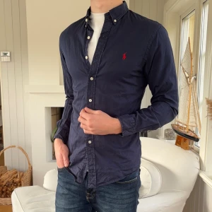 Mörkblå skjorta från Ralph Lauren - Snygg mörkblå skjorta från Ralph Lauren i slim fit med klassisk button-down krage och vit knappstängning. Materialet är lätt bomullstwill och har den ikoniska röda Polo-loggan broderad på bröstet. Perfekt för dig som gillar stilrena och tidlösa plagg.
