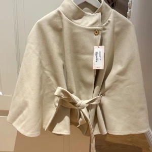 Beige poncho från Nelly  - Helt ny o aldrig använd! Slutsåld på Nelly! Köpt på Drottninggatan i Sthlm men ångrat färg! Stilren beige ponchojacka från Nelly med hög krage, knäppning vid halsen och knytskärp i midjan. Jackan har en mjuk, slät yta och en luftig, svepande passform som ger en cool och avslappnad look.