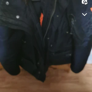 Mörkblå Parajumpers vinterjacka med päls - Säljer en mörkblå Parajumpers vinterjacka i modellen Masterpiece Series. Jackan har huva med pälsdetalj, dragkedja och knappar framtill samt flera fickor. Insidan har orange detaljer och broderade märken. Perfekt för kalla dagar. Pris kan diskuteras vid snabb affär 