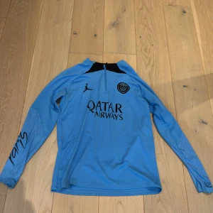 Blå PSG fotbollströja Jordan - Säljer en blå långärmad PSG fotbollströja från Jordan med svart detalj vid halsen och halv dragkedja. Tröjan har tryck med Qatar Airways och PSG-logga på bröstet samt 'Paris' på ena ärmen. Tillverkad i lätt, ventilerande syntetmaterial.