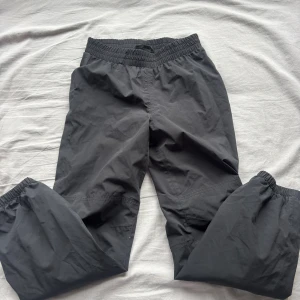 Svarta täckbyxor från Neomondo Junior - Svarta täckbyxor från Neomondo med elastisk midja och diskret logga på benet. Byxorna har ett enkelt, rakt snitt och är gjorda i ett slitstarkt syntetmaterial med meshfoder på insidan. Perfekta för kalla dagar och utomhusaktiviteter.