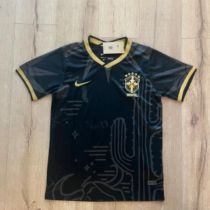 Svart Brasilien fotbollströja Nike M - Snygg svart Brasilien fotbollströja från Nike med guldiga detaljer och broderat CBF-emblem på bröstet. Tröjan har korta ärmar, V-ringad krage med guldkant och subtila grafiska mönster i tyget. Tillverkad i lätt och ventilerande Dri-Fit material.