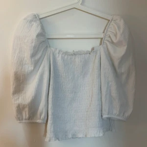 Vit blus med puffärmar XS - Säljer en söt vit blus i storlek XS med puffiga trekvartsärmar och smockad struktur. Toppen har en fyrkantig halsringning och små volangdetaljer vid kanten. Perfekt för dig som gillar romantisk och trendig stil.