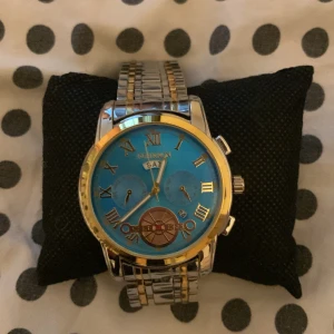 Blå och guld klocka  - Snygg klocka med blå urtavla och romerska siffror i guld. Armbandet är i silver och guldtonad metall med länkdesign. Klockan har flera små urtavlor och detaljer i guld och brons. Perfekt för dig som gillar att sticka ut med accessoarer.