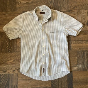 Rutig kortärmad skjorta Ben Sherman - Sjukt nice kortärmad skjorta från Ben Sherman i rutigt mönster med beige, bruna och vita toner. Klassisk krage, knäppning framtill och broderad logga på bröstet. Perfekt för dig som gillar en clean och tidlös stil.