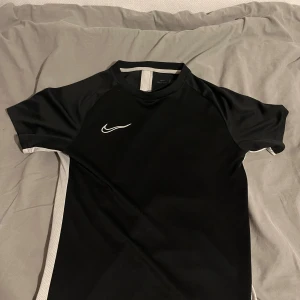Svart Nike Dri-Fit träningströja S - Nike Dri-Fit träningströja i svart med vita detaljer längs sidorna och vid halsen. Tröjan har korta ärmar och är tillverkad i ett lätt, ventilerande funktionsmaterial som håller dig torr under träningen. Snygg och sportig design med Nike-logga på bröstet.