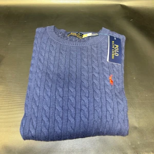 Marinblå kabelstickad tröja  - Kabelstickad marinblå tröja med rund halsringning och klassisk röd broderad logga på bröstet. Tröjan är i bomull och har en tidlös, stilren look som passar perfekt till jeans eller chinos.