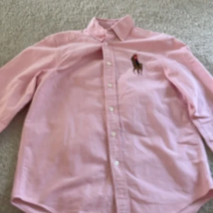 Rosa skjorta från Ralph Lauren M - Klassisk rosa skjorta från Ralph Lauren med broderad polospelare på bröstet. Skjortan har knappar framtill, krage och långa ärmar. Tillverkad i mjuk bomull och passar perfekt för en stilren look.