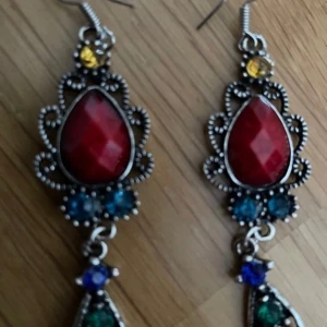 Färgglada droppformade örhängen - Unika örhängen med droppformad röd sten i centrum, omgivna av detaljerad silverfärgad metall. Dekorerade med små stenar i blått, grönt, gult och rosa. Ca 6 cm långa, perfekta för dig som gillar färg och statement-smycken.