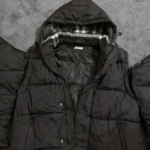 Svart pufferjacka från Burberry - Svart pufferjacka från Burberry med stor logotyp på bröstet och klassiskt rutigt foder i huvan. Jackan har dragkedja, tryckknappar och två fickor framtill. Perfekt för kalla dagar och har en riktigt clean och modern look.